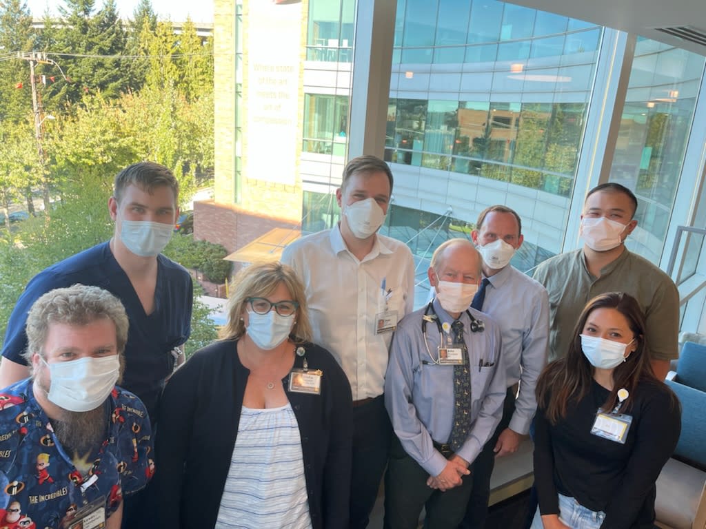 Eight people wearing masks stand together in front of a window. A building with a glass facade is visible in the background, with trees on the left.