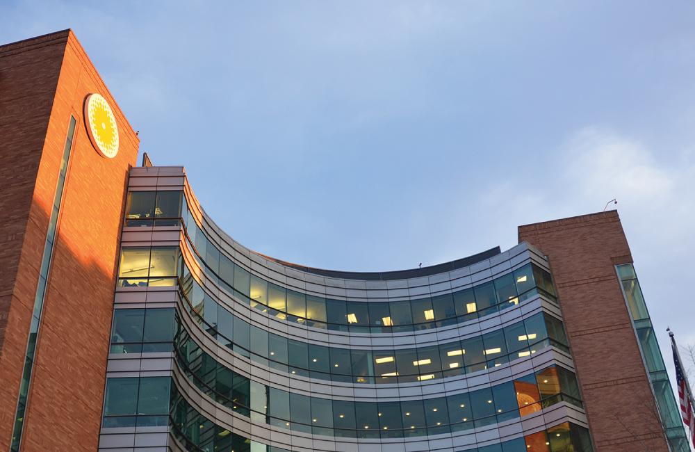 Large brick medical building at sunset with curved glass facade and lights on inside.