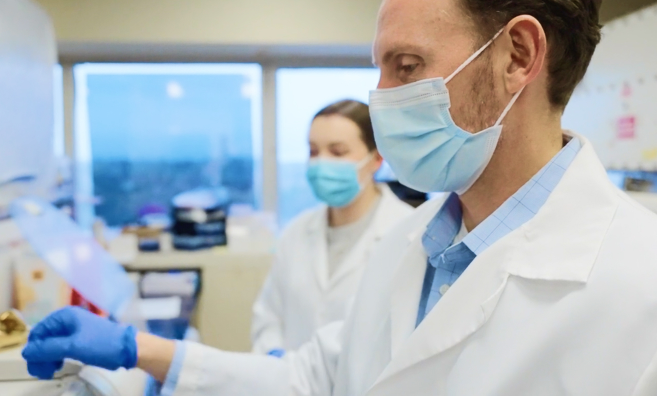 Dr. Blosser and a colleague, wearing lab coats and gloves, conduct research in a lab.