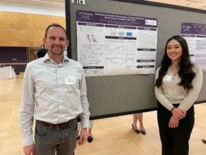 Dr. Blosser and Regina Tsay smiling, standing on either side of a research poster in a conference hall.